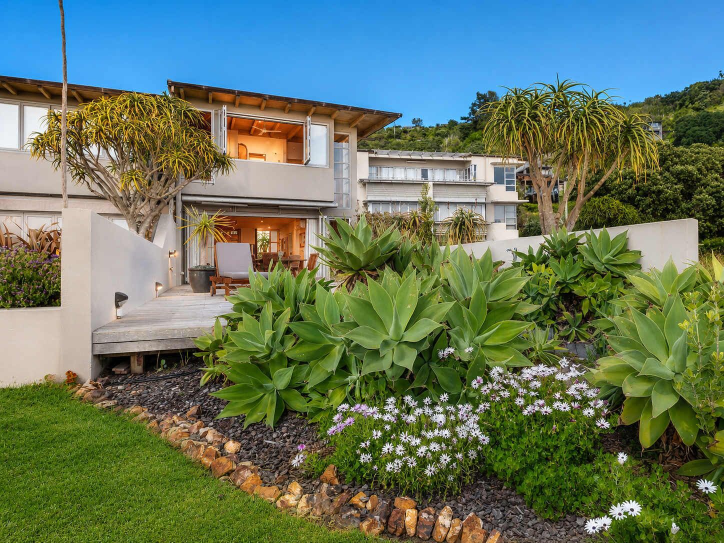 A modern two-story house with a patio, surrounded by lush greenery and flowering plants.