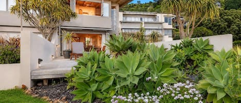 Una casa moderna de dos pisos con un patio, rodeada de exuberante vegetación y plantas florecientes.