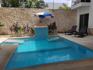 A swimming pool with a stone wall, a blue and white umbrella, and two lounge chairs. - ney Tulum apartments (Tulum)