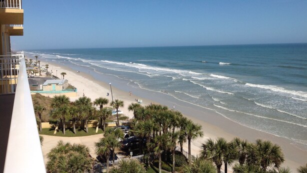 A beachfront view from a balcony overlooking a sandy beach with palm trees and a clear blue sky.