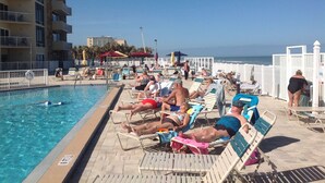 A poolside area with sun loungers and a pool, surrounded by a white fence and a building with balconies.