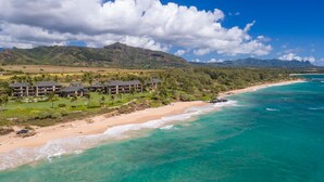 Un complejo turístico costero con propiedades frente a la playa, un campo de golf y terreno montañoso al fondo.