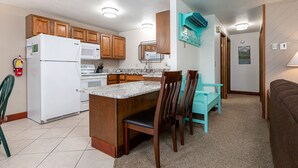 A compact kitchen with wooden cabinets, a white refrigerator, a microwave, and a stove. There is a small island with chairs and a teal wall-mounted air conditioning unit.
