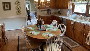 A dining area with a round table, white chairs, a chandelier, wooden cabinets, and a window with curtains. - Beekeeper's House (Buena Vista)