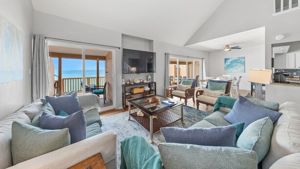 A spacious living room with a large sofa, armchairs, and a coffee table. There is a television mounted on the wall, and a balcony with a view of the ocean.