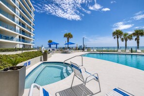 A pool area with a clear view of the ocean, a building with balconies, and lounge chairs. - Oceanfront Corner Condo with Wraparound Balcony Heated Pool and Hot Tub (New Smyrna Beach)