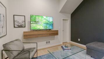 A modern living room with a flat-screen TV mounted on a wooden console, a grey sofa, a glass coffee table, and a framed picture on the wall.