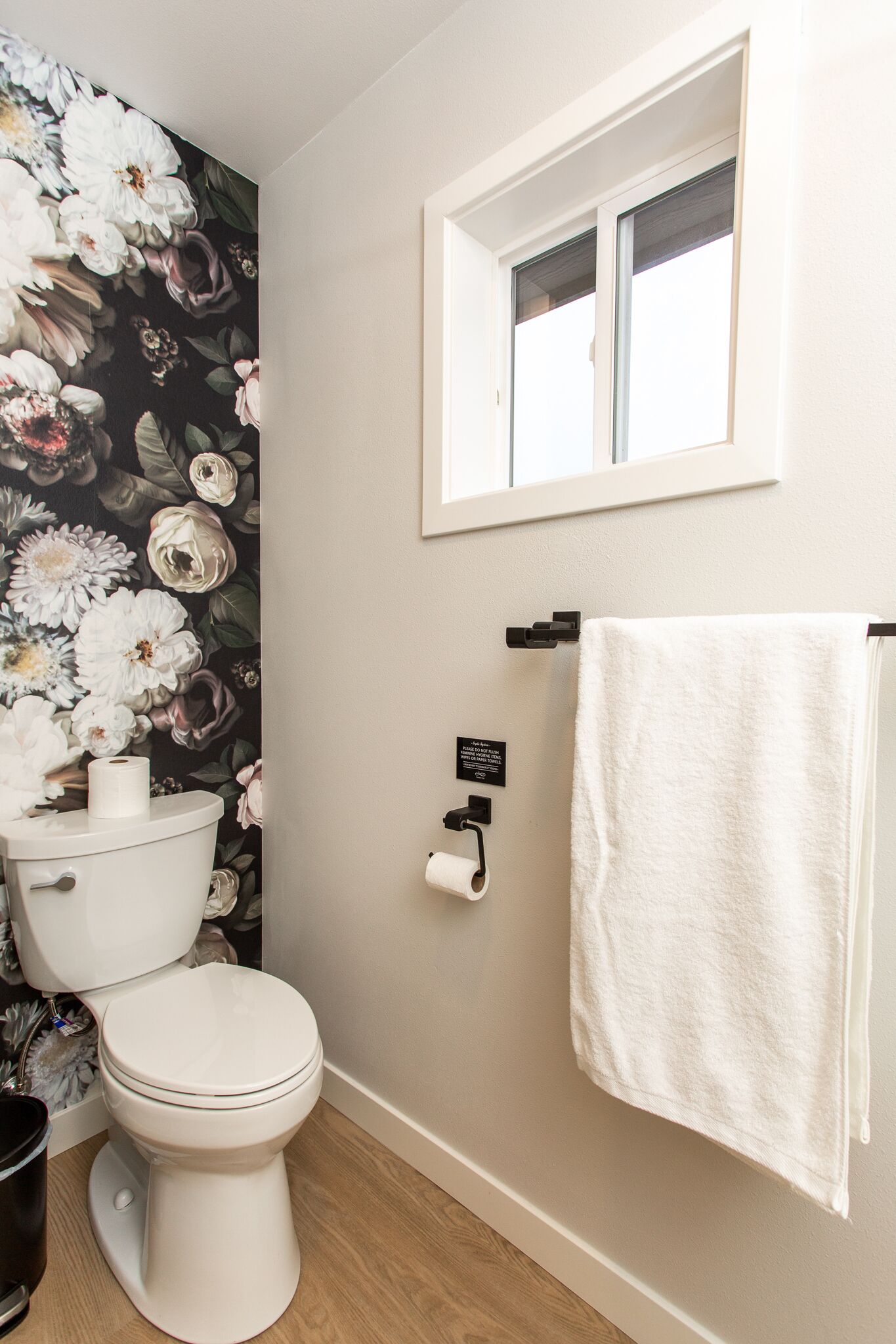 Ein Badezimmer mit Toilette, Toilettenpapier, einem Handtuch auf einem Haken und einem Fenster über der Toilette.