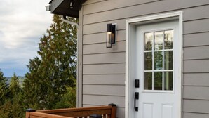 A house with a white front door, a small porch with a wooden bench, and a wall-mounted light fixture. - Stunning Views from this Modern Hood Canal Retreat (Union)