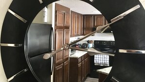 A large wall clock with a mirror, reflecting a kitchen area with wooden cabinets and a stove.