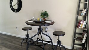 A dining area with a round table, two stools, a potted plant, and a bookshelf against the wall.