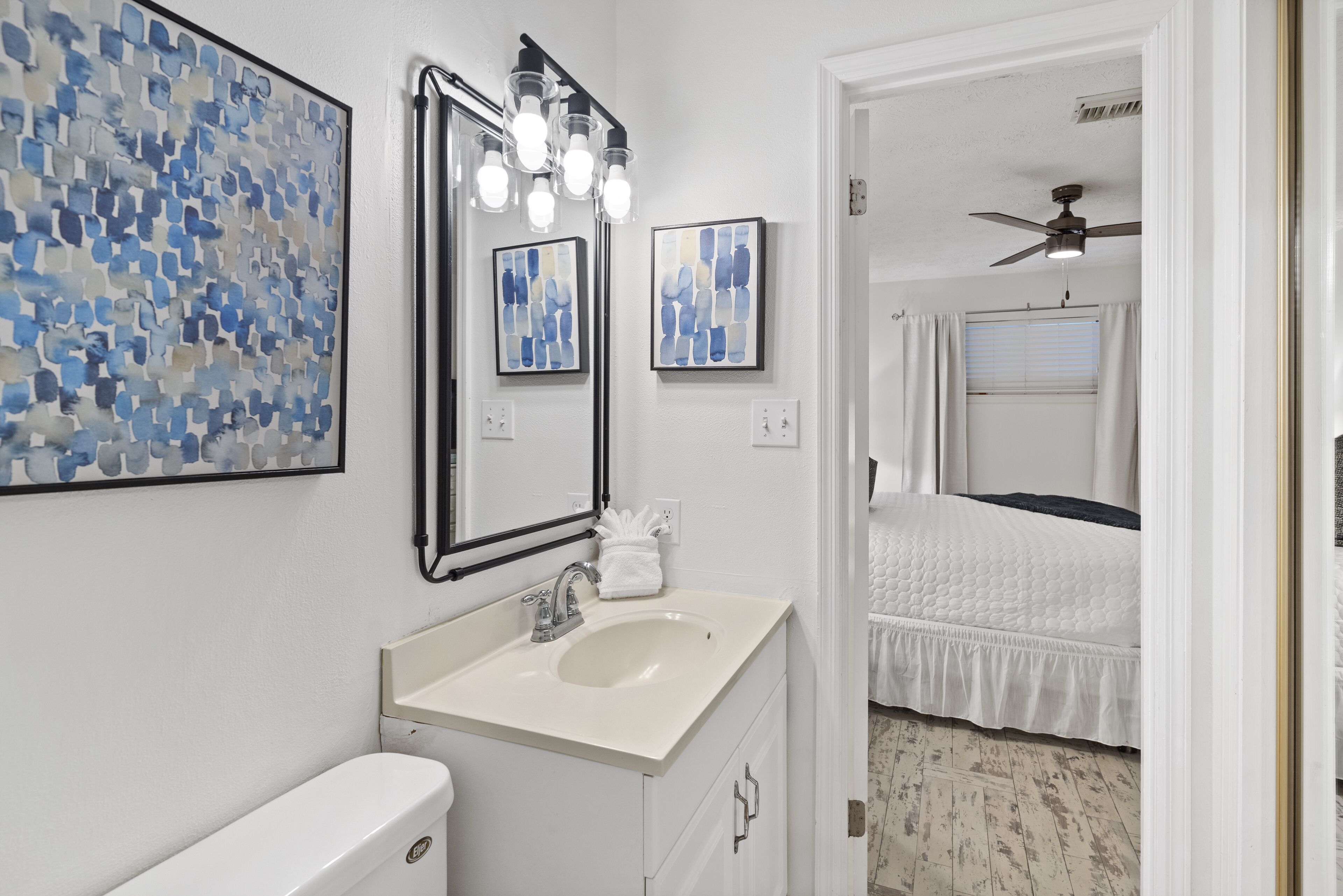 Une salle de bain avec un lavabo blanc, des toilettes et un miroir avec éclairage. Deux œuvres d’art encadrées sont accrochées au mur.