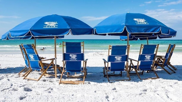 A beach with four blue beach chairs and two large blue umbrellas with the Coral Reef Club logo. - Location Location Location!! (Destin)