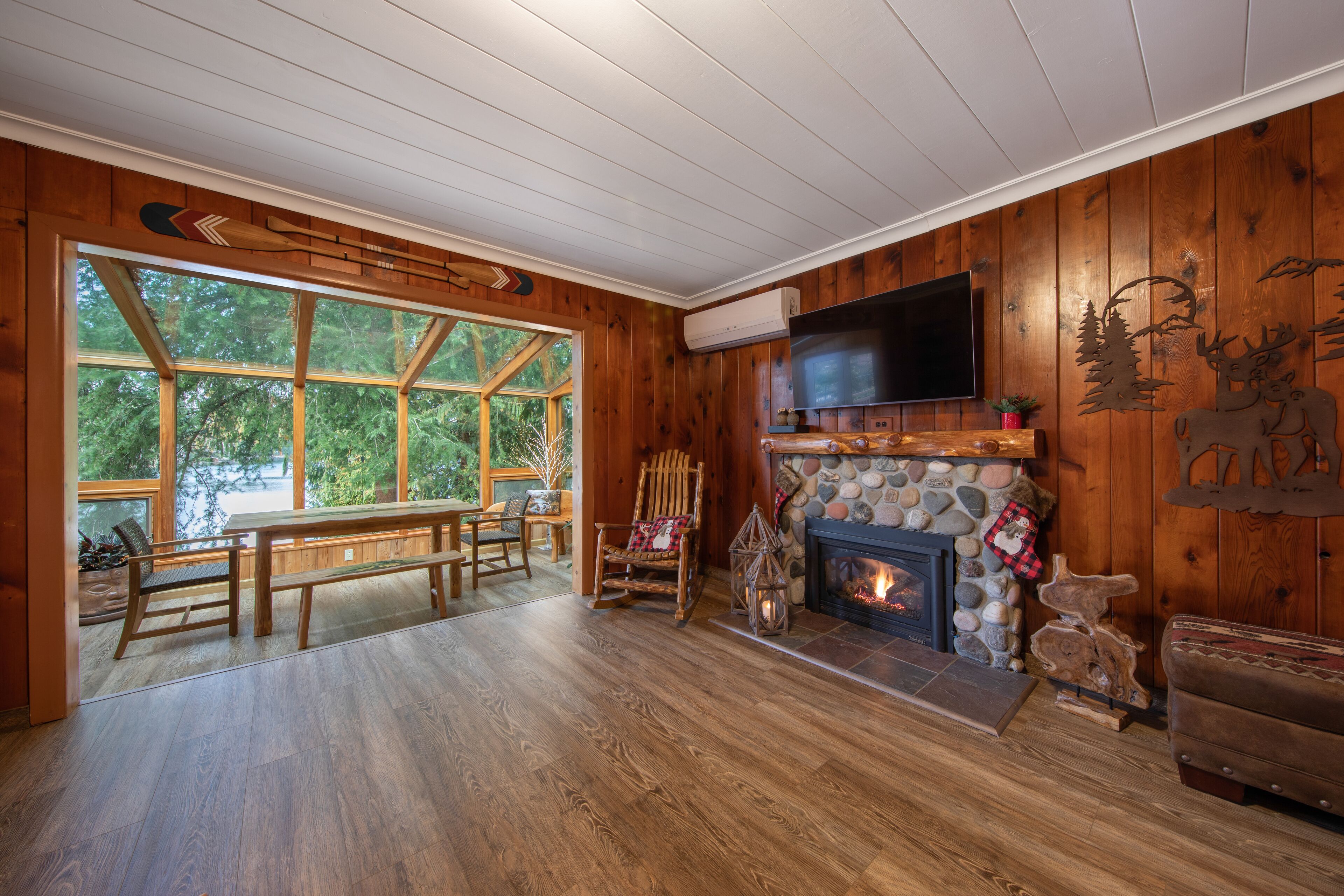 A cozy cabin interior with a stone fireplace, wooden walls, and a large window overlooking a forest.