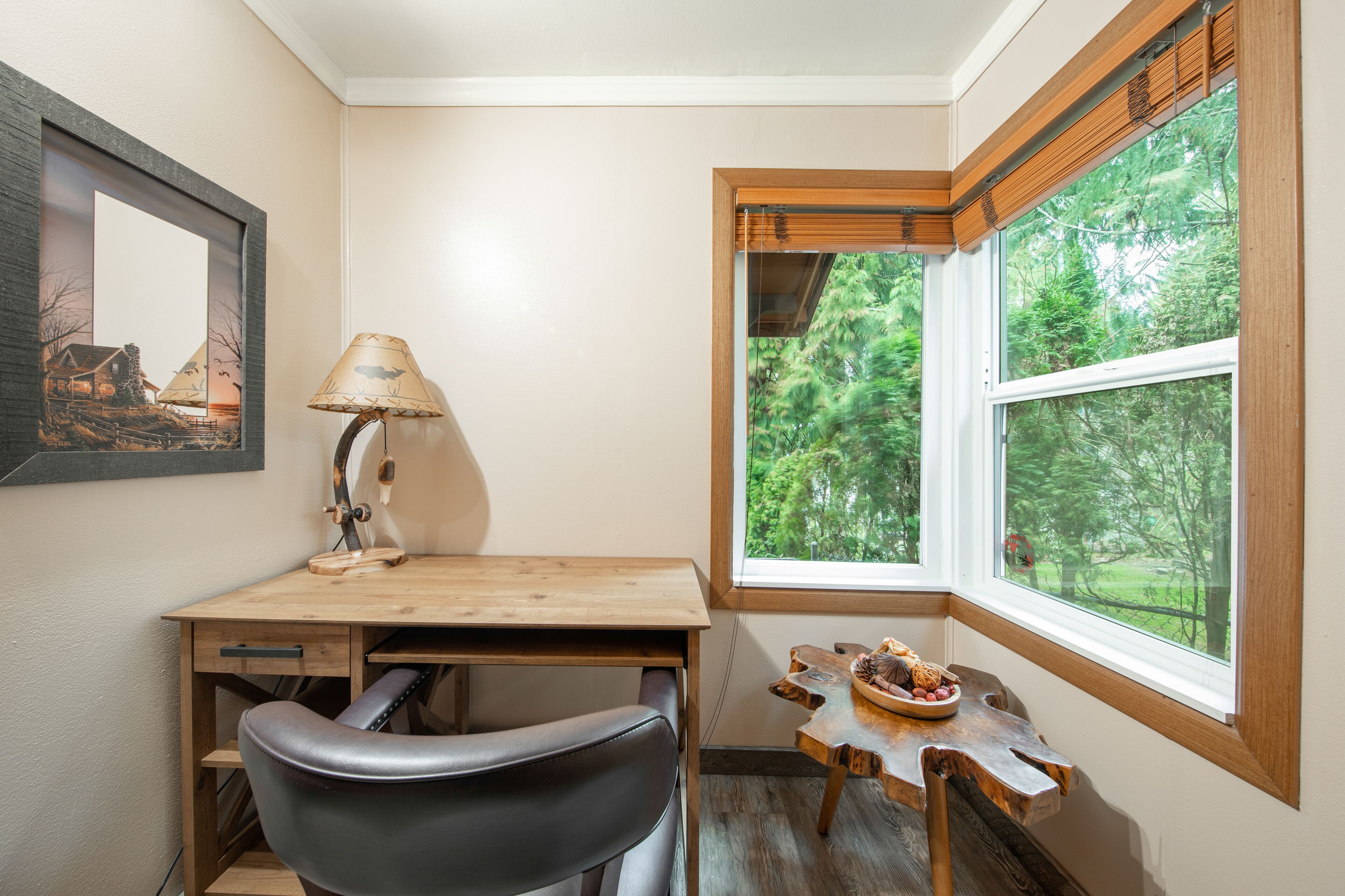A corner room with a wooden desk, a black chair, a lamp, a small table with a plate of food, and a large window with a view of trees.