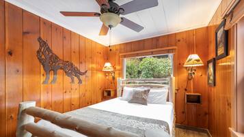 A wooden-paneled bedroom with a bed, two wall-mounted lamps, a ceiling fan, and a window with a view of greenery.