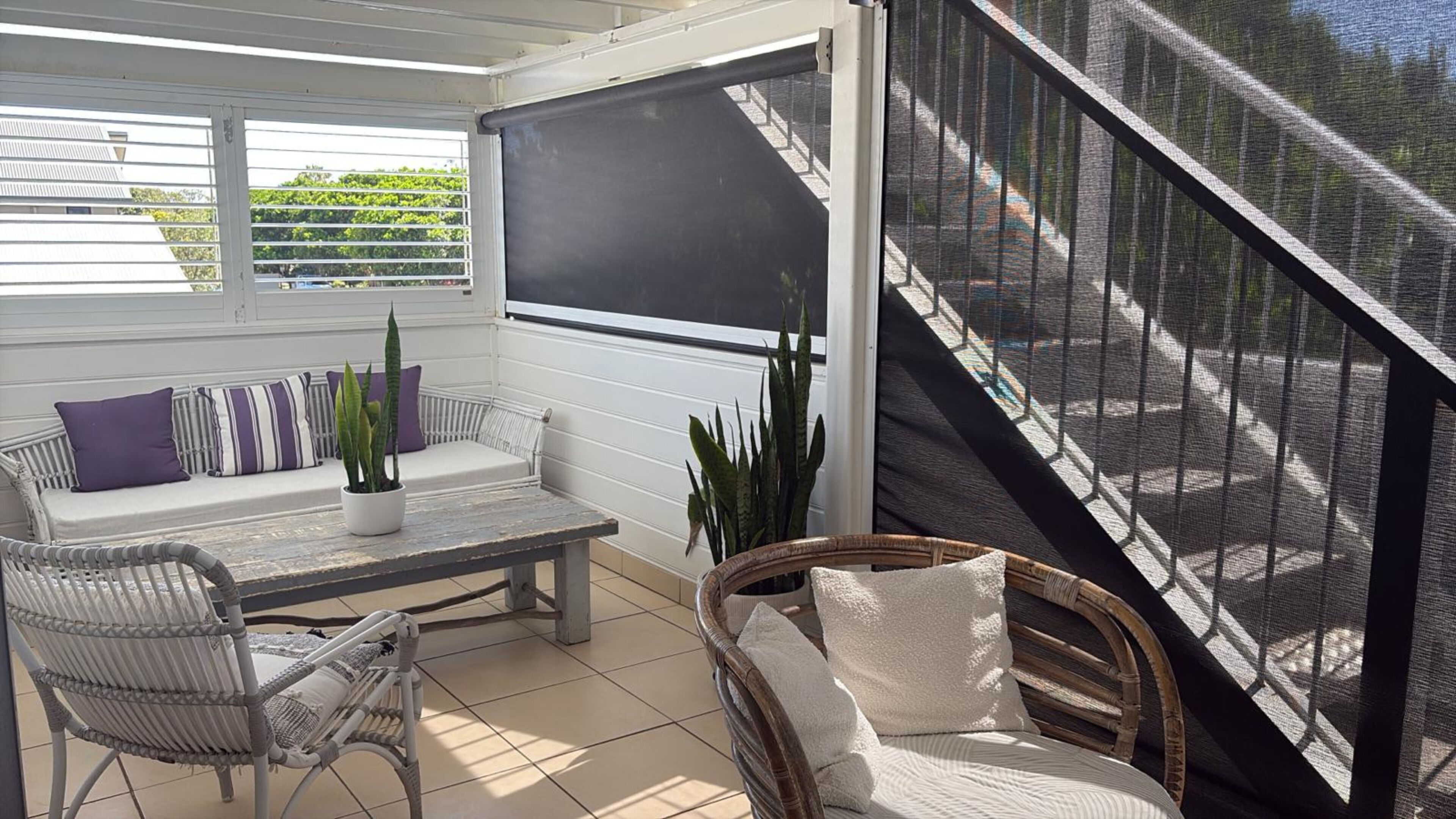 Une terrasse ensoleillée avec un canapé blanc, un fauteuil en osier, une petite table et un grand écran.