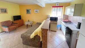 Un espacio habitable compacto con sofá, televisión, una mesita con una planta, y una cocina con microondas, estufa y fregadero.