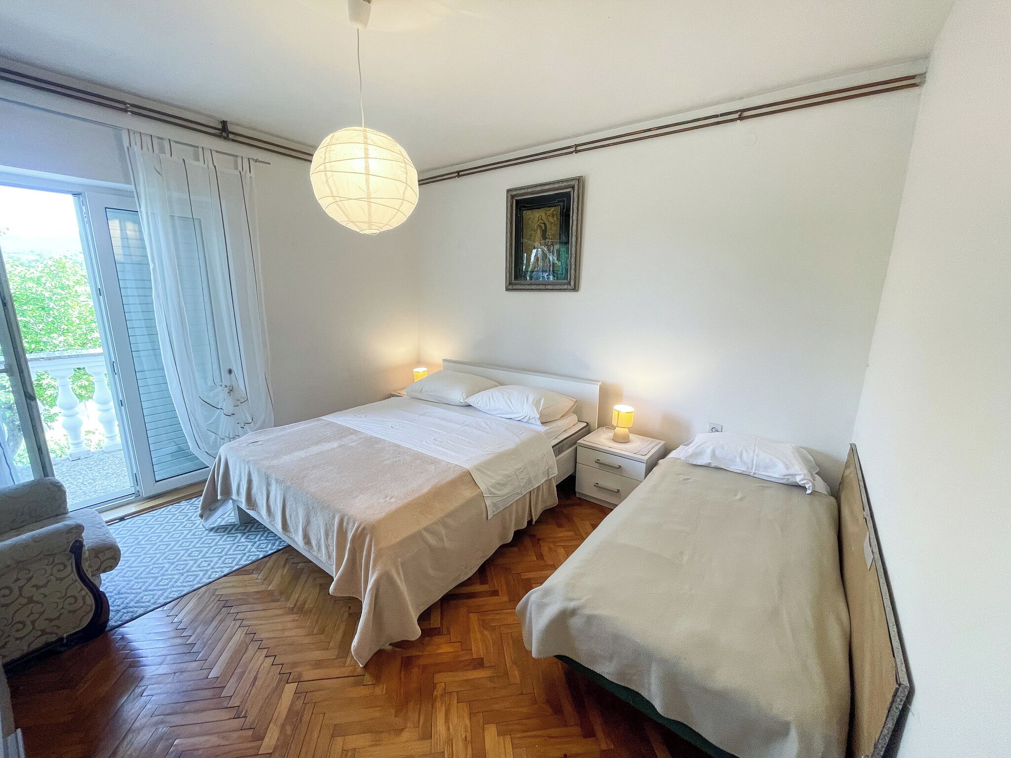 A bedroom with two beds, a nightstand, a lamp, a painting, and a view of the outdoors.