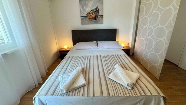 A neatly made bed with striped bedding and rolled towels, a painting on the wall, and a patterned curtain. - Holiday apartment with balcony and air conditioning (Jadranovo)