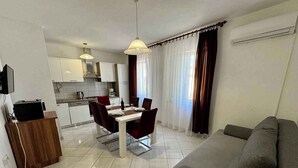 A modern living room with a grey sofa, a dining area with red chairs, a kitchen with white cabinets, and a TV mounted on the wall. - Holiday apartment with balcony and air conditioning (Jadranovo)