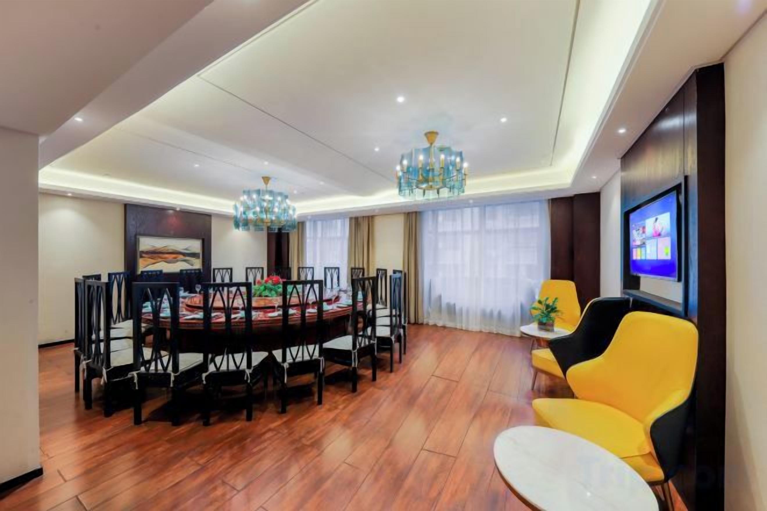 A dining area with a round table surrounded by chairs, a television on the wall, and a chandelier.