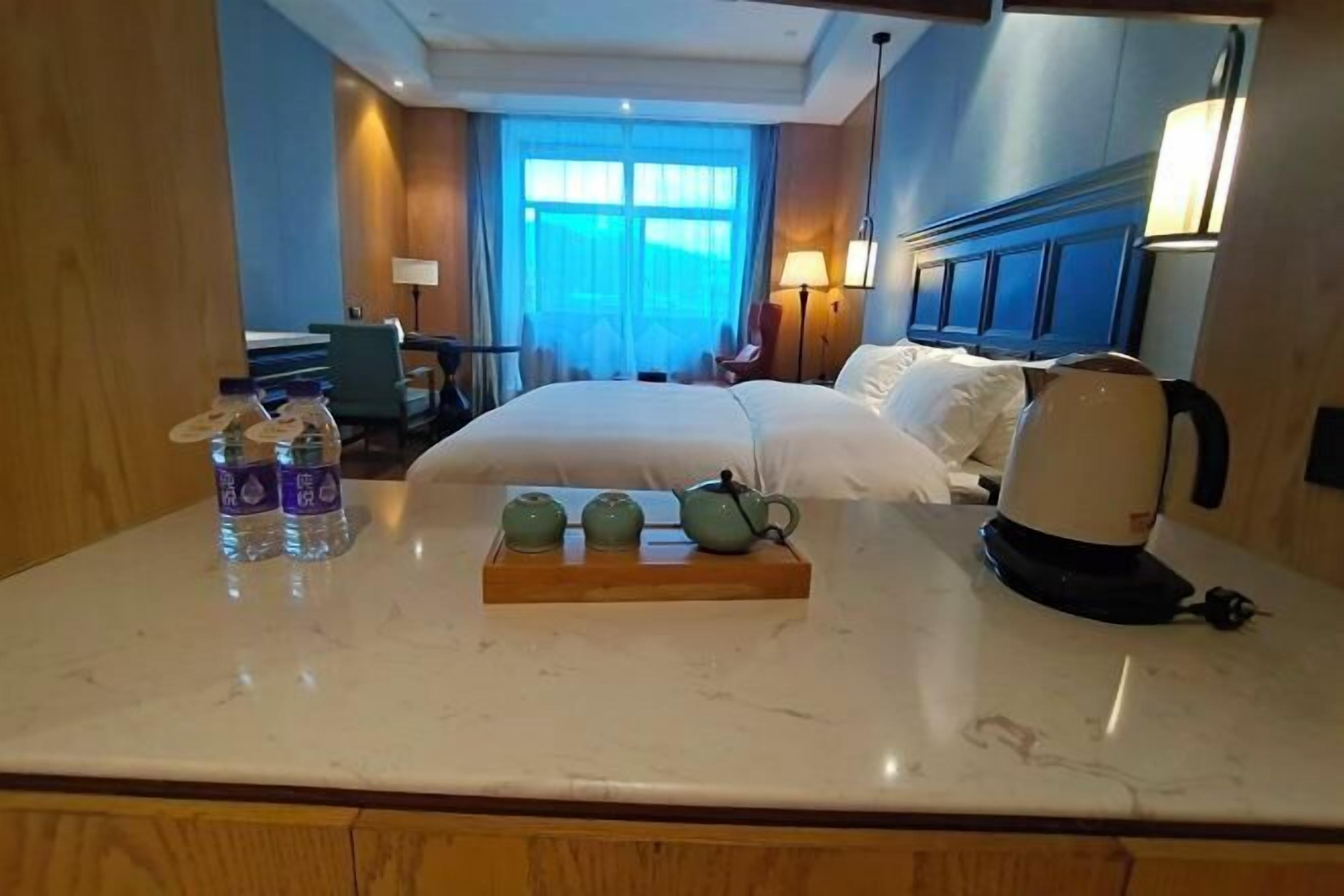 A hotel room with a bed, bedside table, lamp, and a view of the outside through a window.