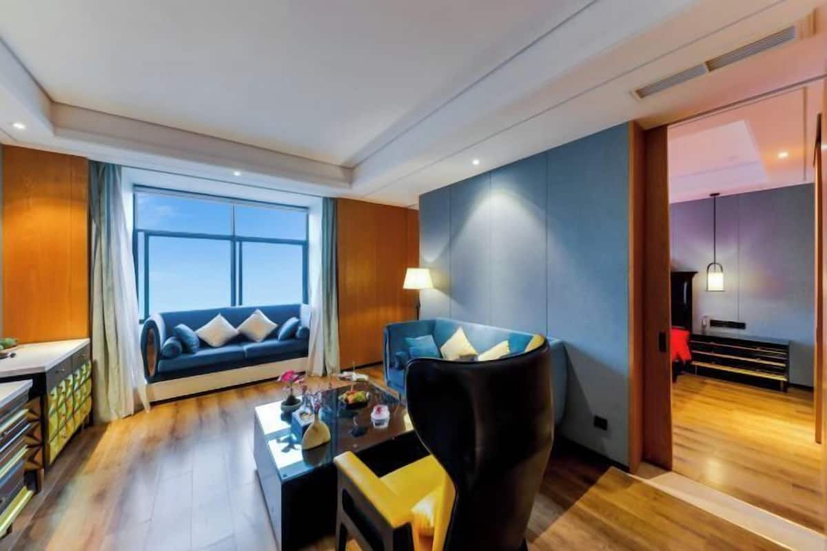 A modern hotel room with a large window, a sofa, a chair, a coffee table, and a desk.