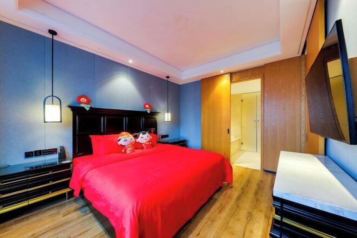 A modern hotel room with a large bed, wooden flooring, a dark headboard, and a flat-screen TV mounted on the wall.