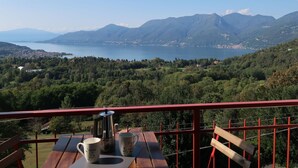Un balcón con una mesa, dos tazas y una cafetera. Se divisa una vista panorámica de un lago y montañas al fondo.