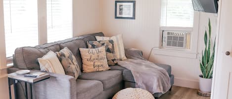 A cozy living room with a grey sectional sofa, a small side table, a woven pouf, and a patterned rug. There are two framed pictures on the wall, a potted plant, and a TV mounted on the wall.