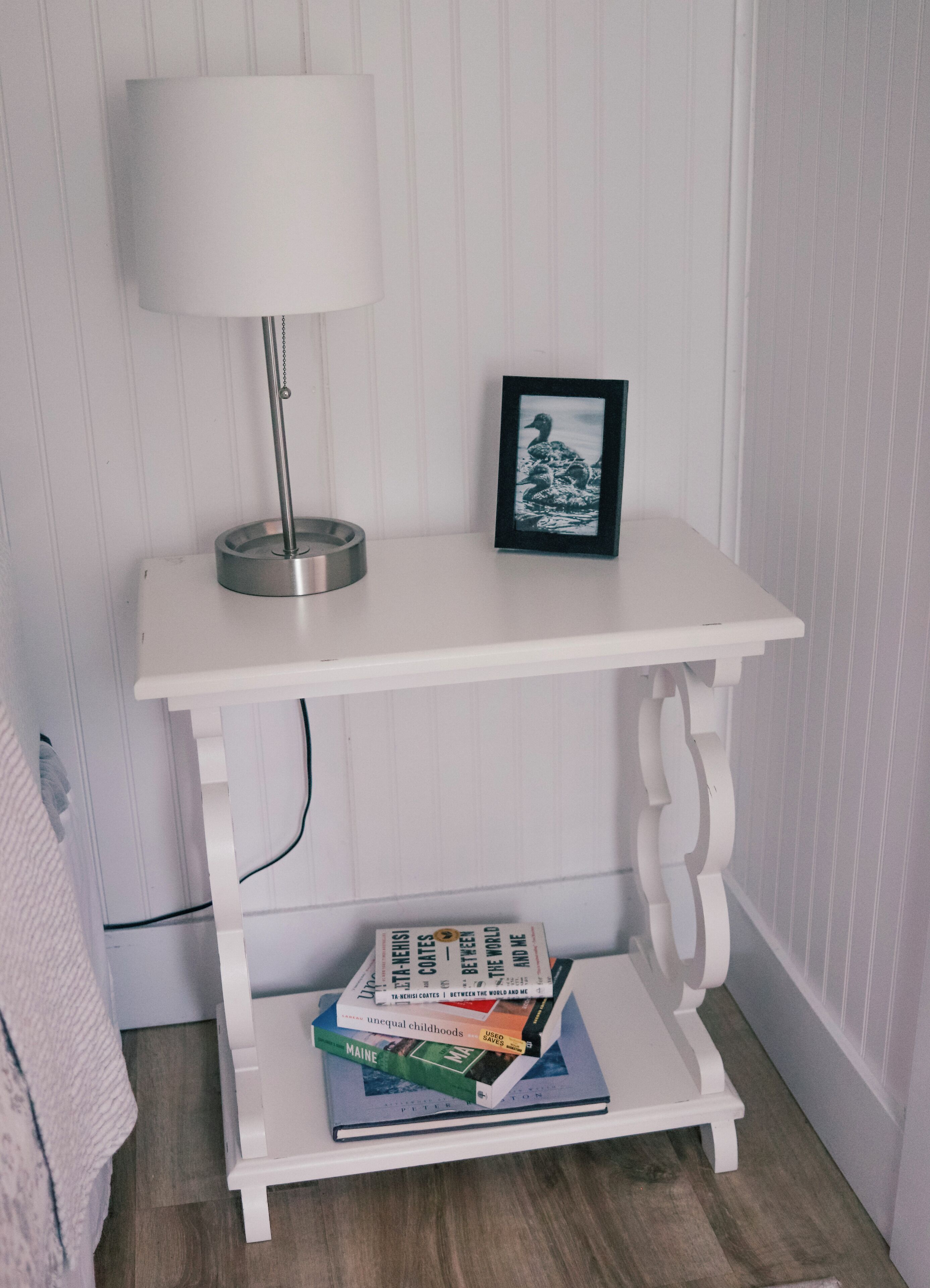 A white console table with a lamp and framed picture, a shelf with books, and a cord on the side.
