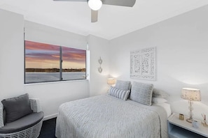 Une chambre à coucher comprenant un lit, une chaise, une table de chevet, une œuvre d’art murale et une fenêtre donnant sur un coucher de soleil.