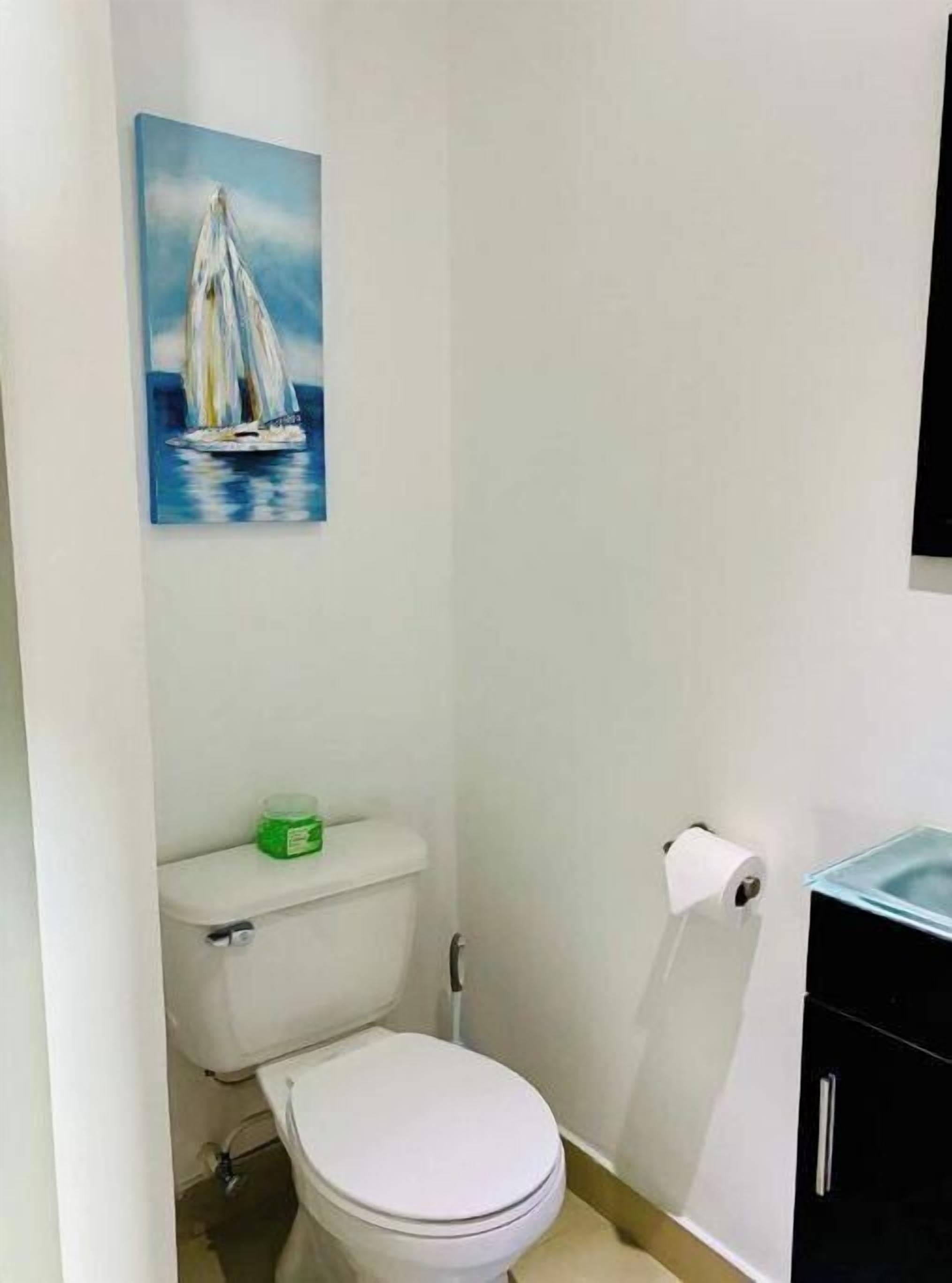 A small bathroom with a toilet, a green container on the tank, a painting of a sailboat, and a black cabinet.