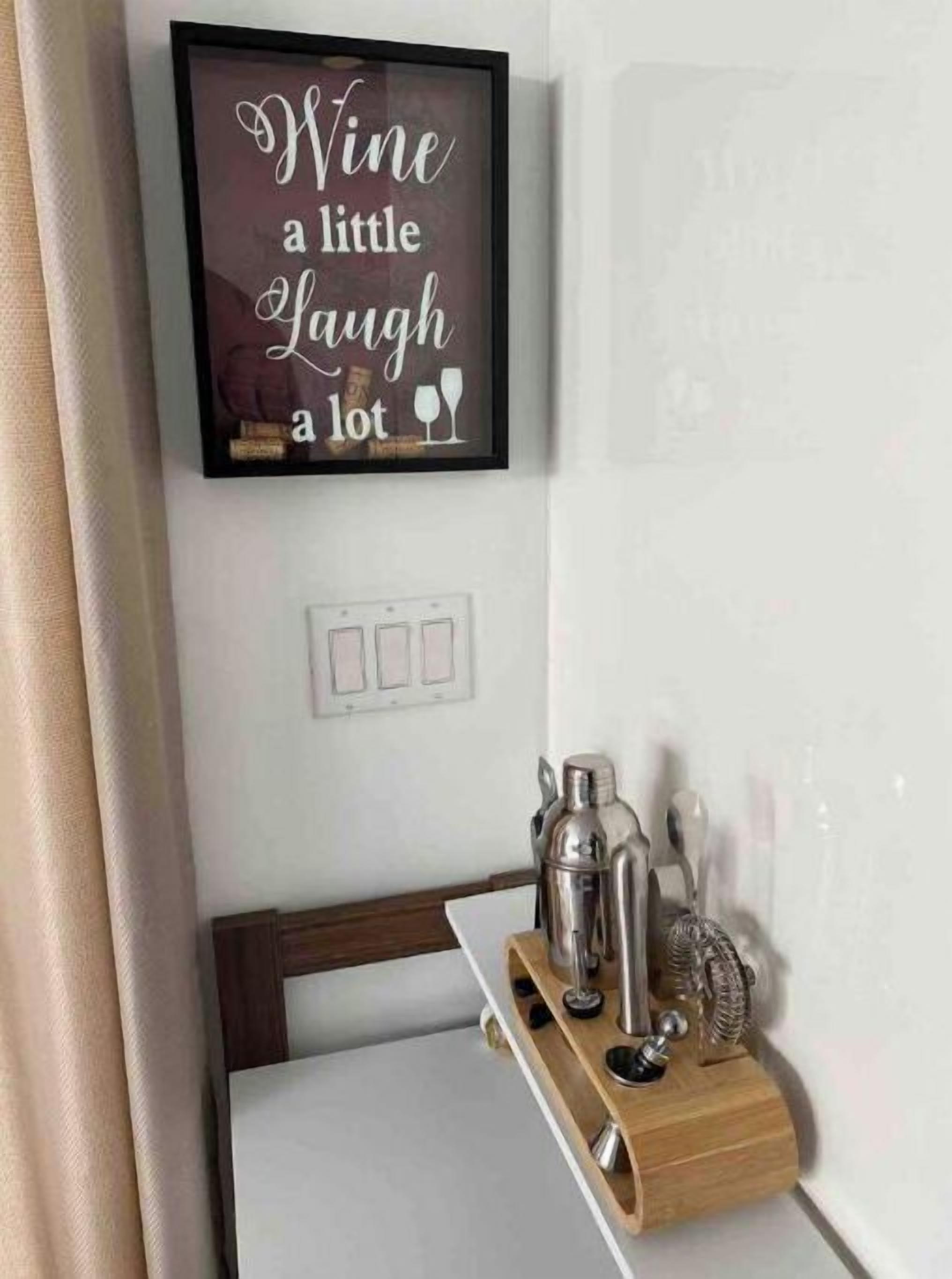 Ein gerahmtes Schild mit der Aufschrift 'Wine a little Laugh a lot' über einem Holzboden mit einem Cocktail-Shaker und weiteren Gegenständen.