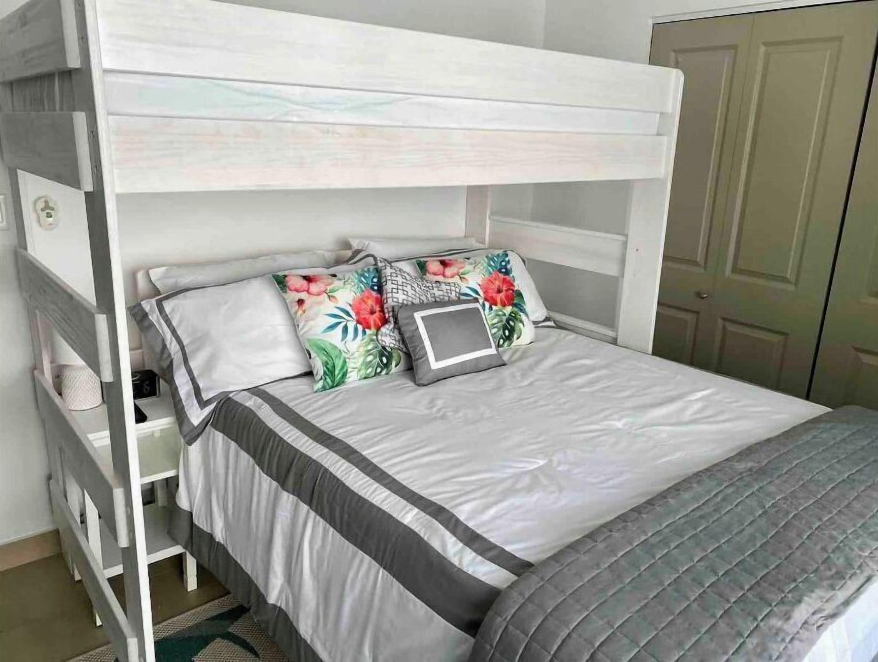 Una litera con estructura blanca, colcha gris y blanca, y almohadas estampadas con motivos florales.