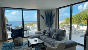 A modern living room with a large sofa, a coffee table, and a view of the sea. - Villa Crystal Clear - Luxury Villa with Private Pool (Sint Maarten)