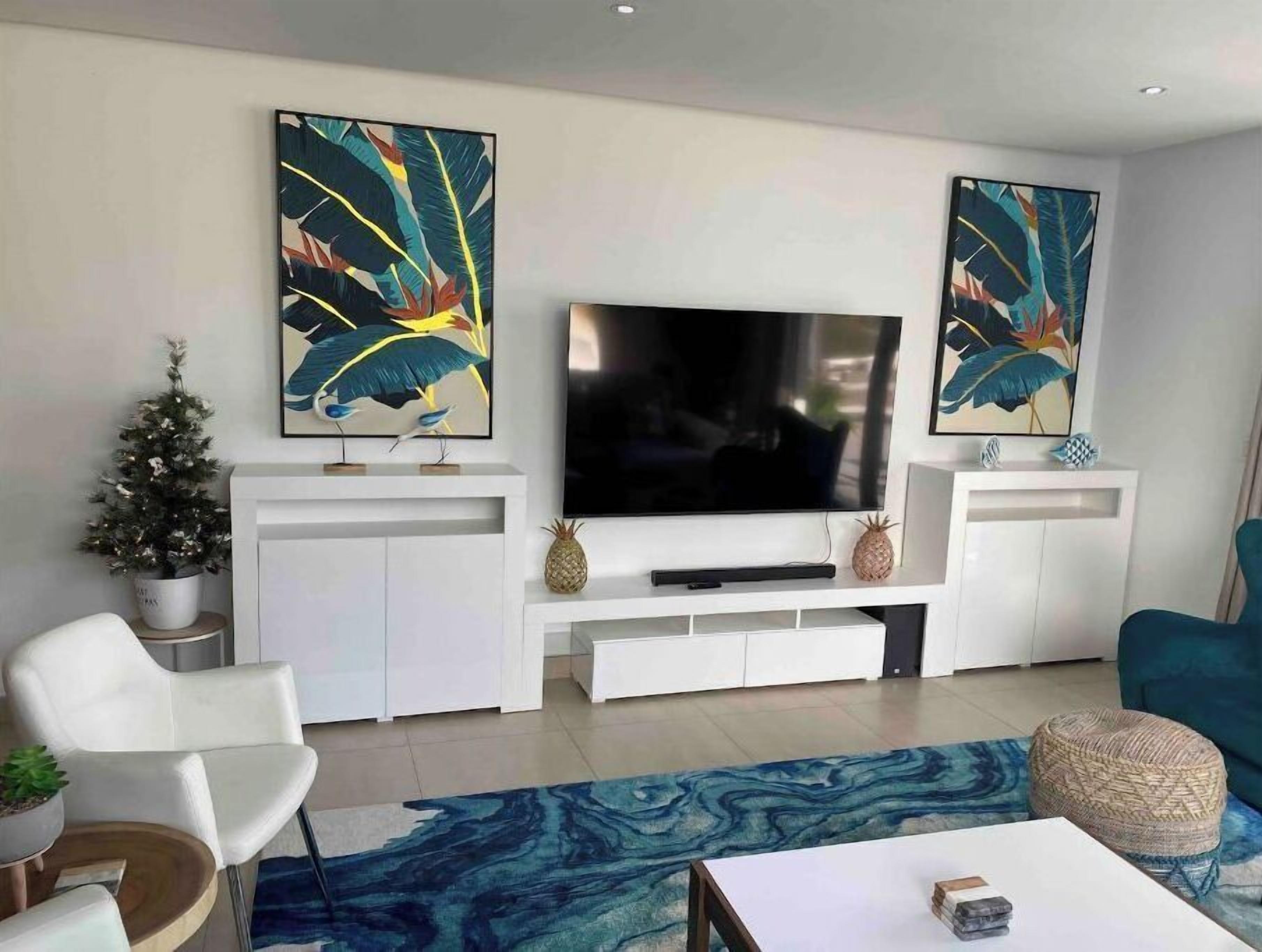 Un salon moderne avec un téléviseur à écran plat, des meubles blancs, des œuvres d’art décoratives, un petit sapin de Noël et un tapis bleu à motifs.