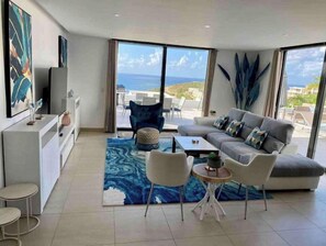 Relax in this spacious living room with a sofa set, coffee table, and two chairs, offering a view of the ocean and coastline.