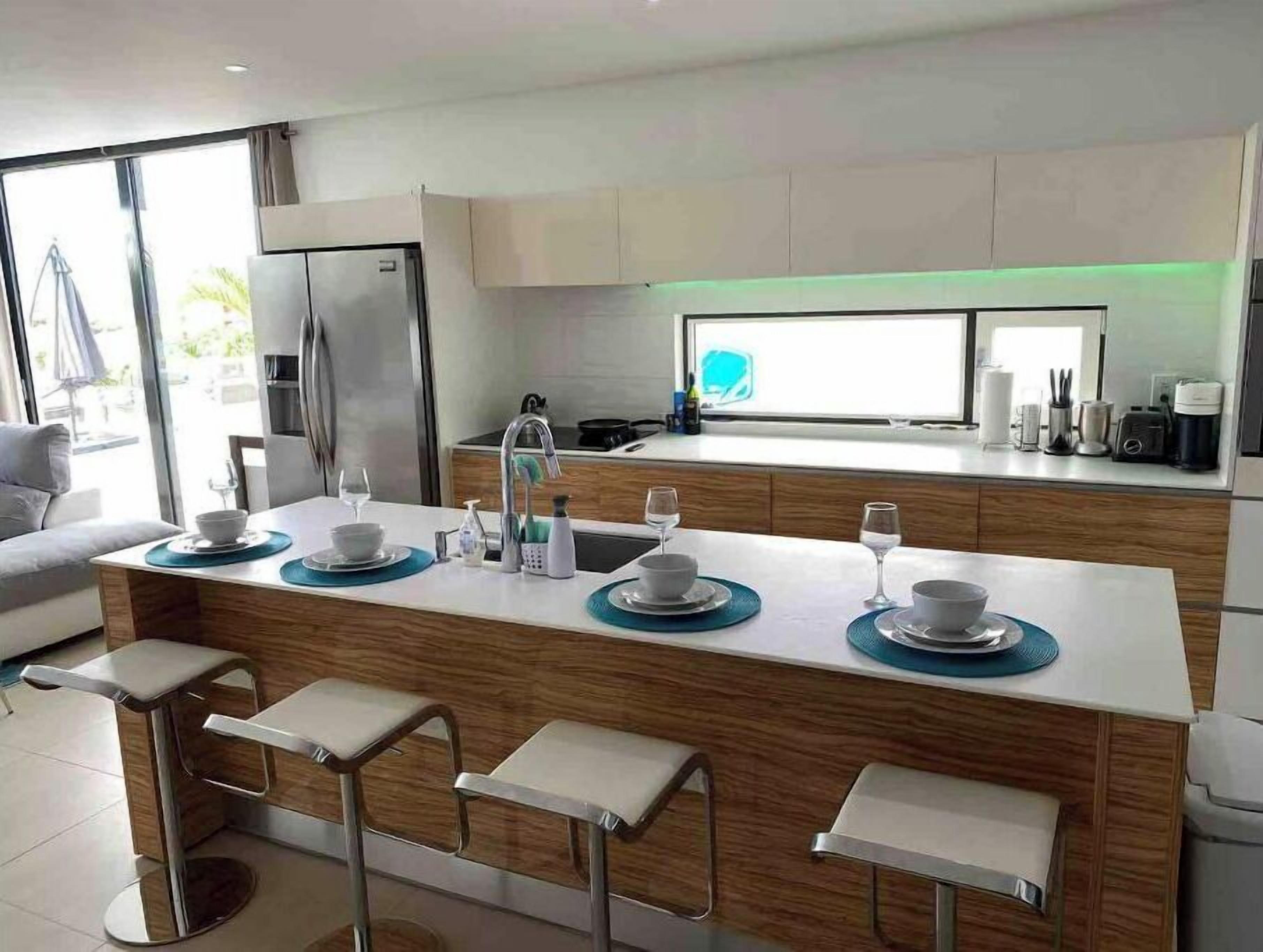 A modern kitchen with a central island, bar stools, a refrigerator, and a large window.