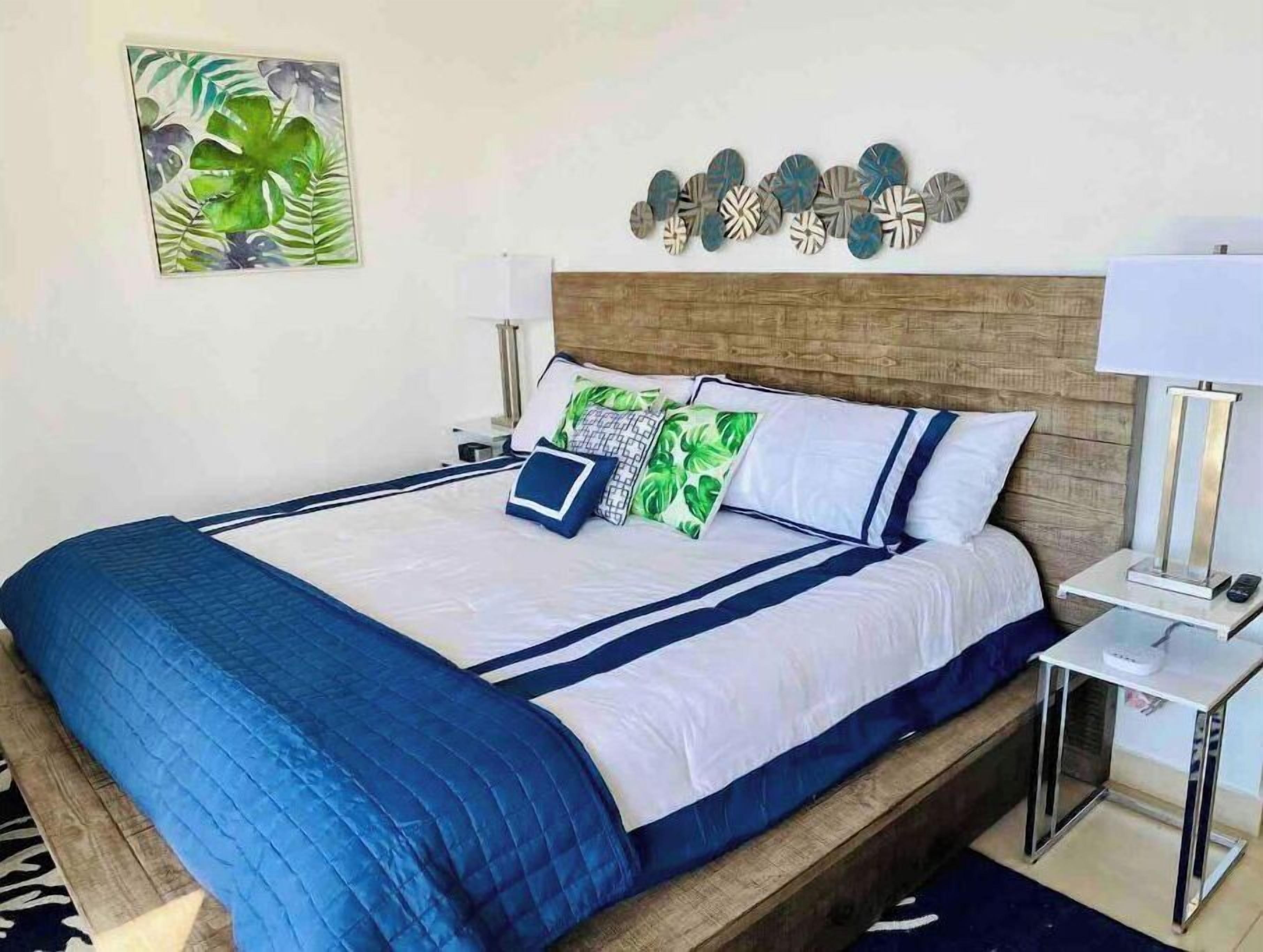 A neatly made bed with a blue quilt, white bedding, and decorative pillows. There is a bedside table with a lamp and a remote control.