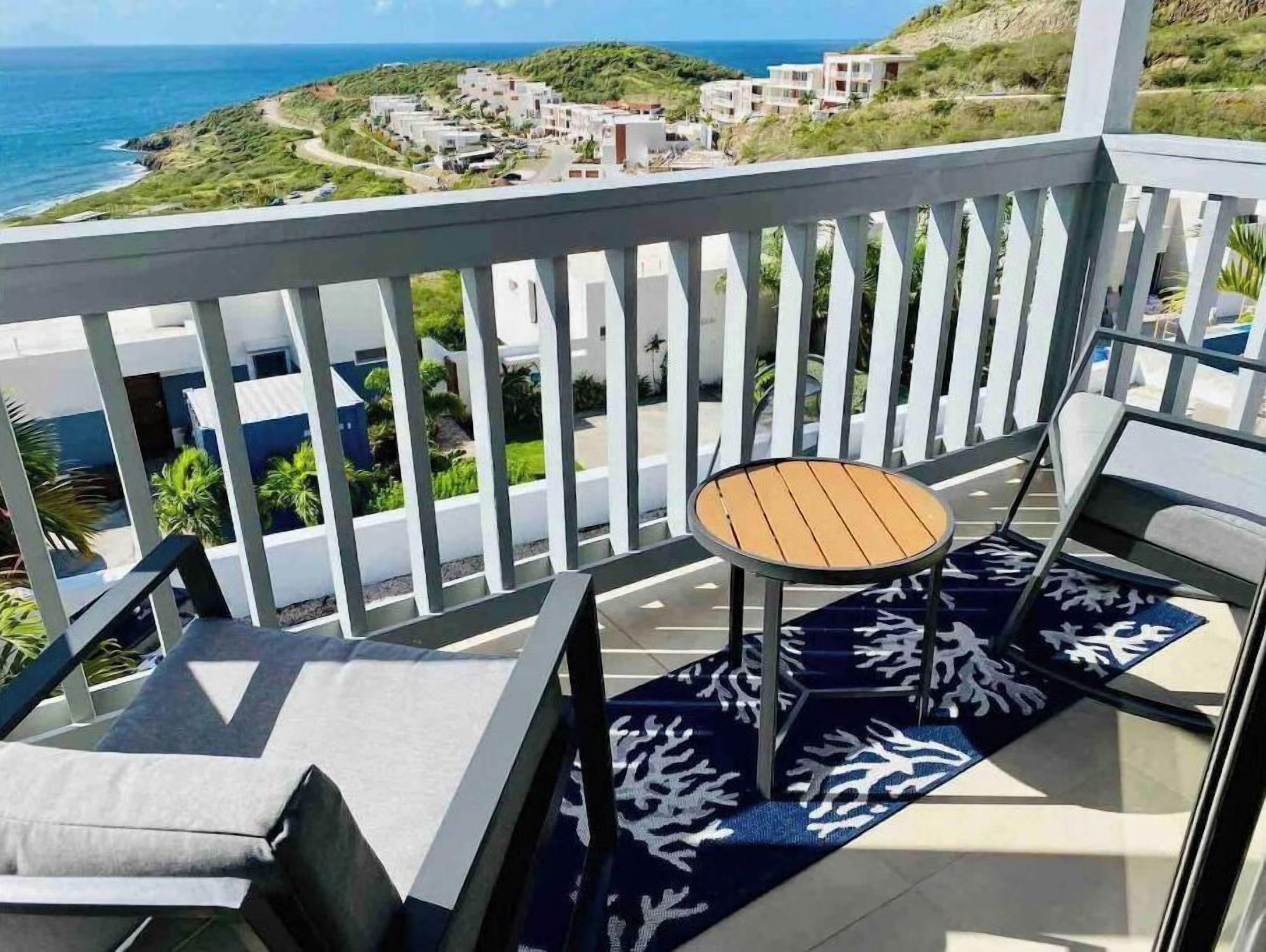 A balcony with a table and chairs, overlooking a coastal area with buildings and greenery.
