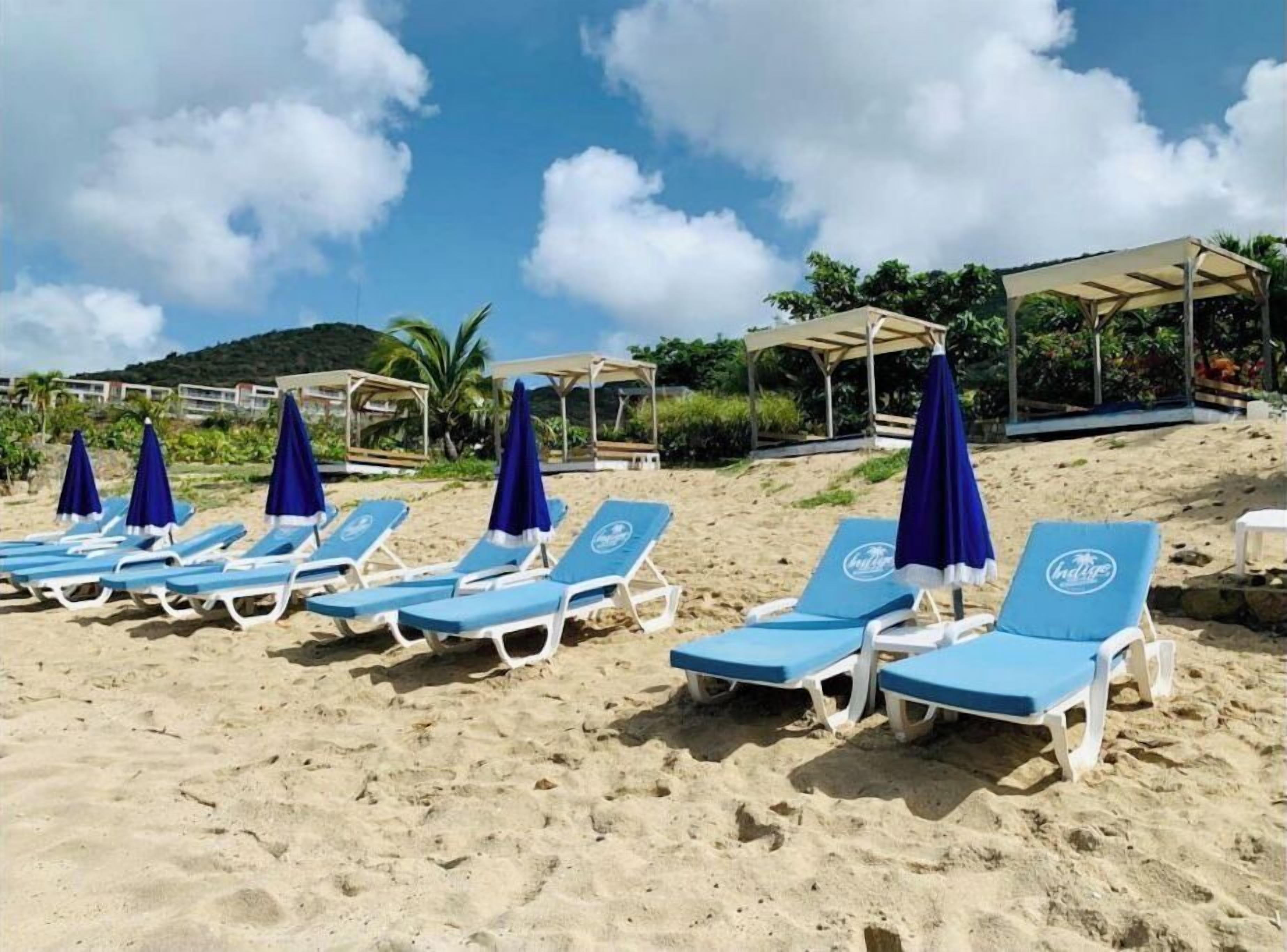 A beach with rows of blue lounge chairs and matching umbrellas, a covered seating area, and a backdrop of greenery and hills.