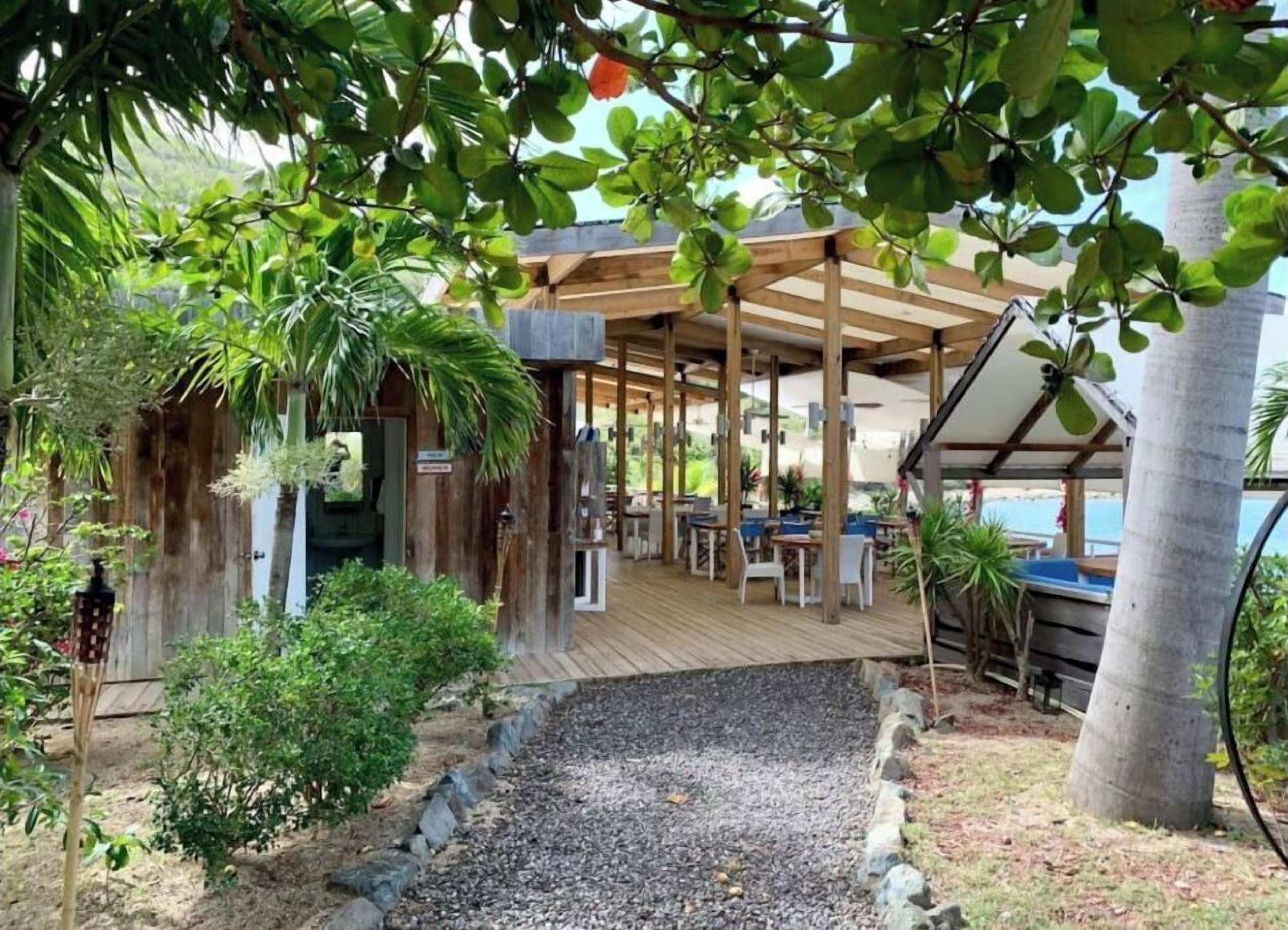 Un espacio al aire libre cubierto con mesas y sillas de madera, rodeado de vegetación y un sendero de grava.