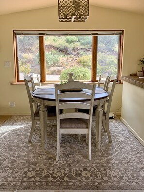 A dining area with a table and chairs, a view of a rocky landscape through large windows, and a patterned rug on the floor.