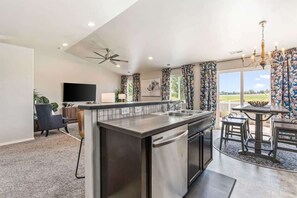A modern kitchen with a central island, a dining area with a table and chairs, a ceiling fan, and a large window with curtains. - Private Wine Country Retreat | South Nampa | Sleeps 10 (Nampa)