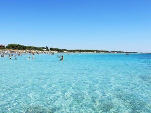 A beach with clear turquoise water, people swimming and sunbathing, and a distant shoreline with buildings and greenery.