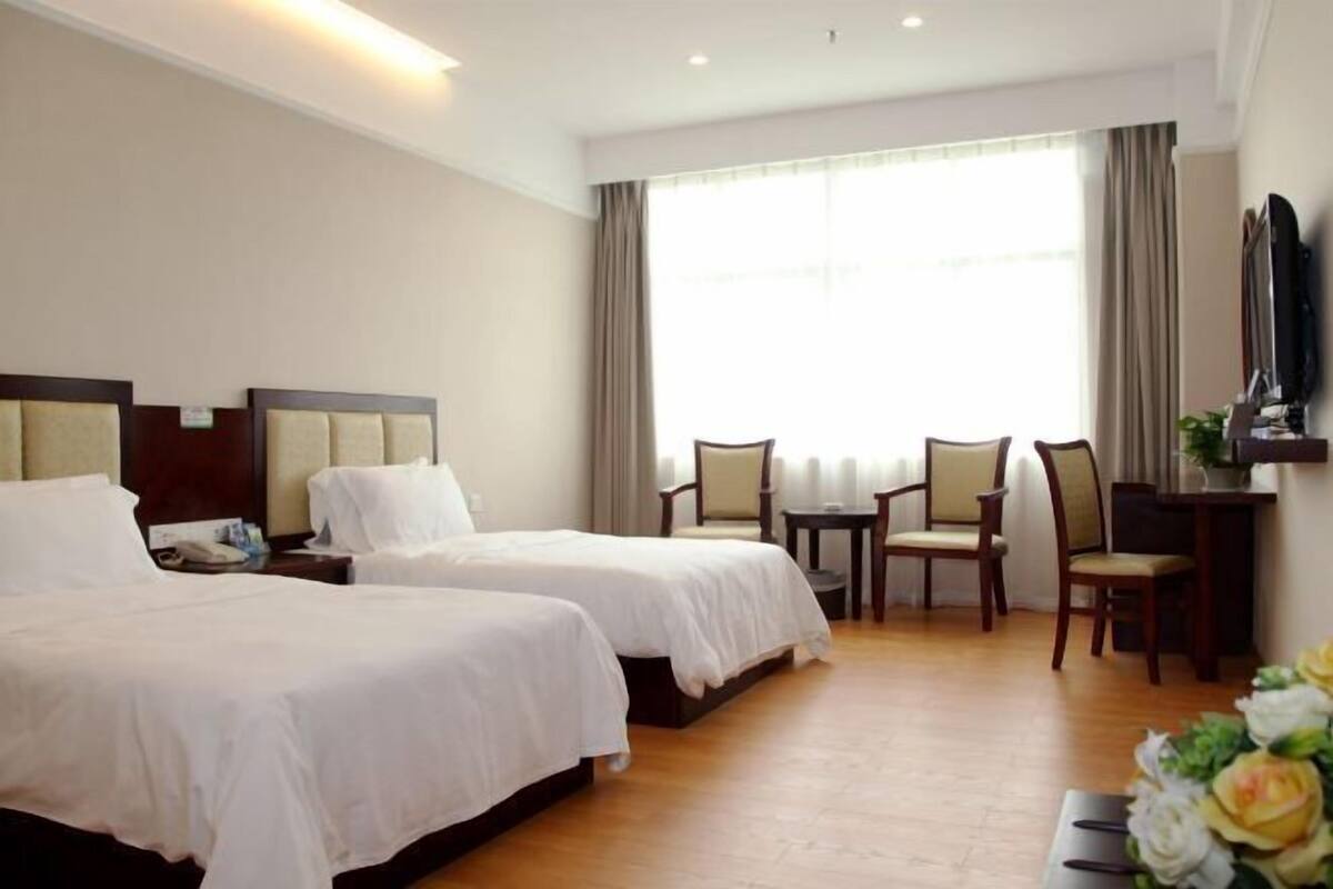 Habitación de hotel con dos camas, un escritorio, una silla, un televisor y un ventanal grande con cortinas.