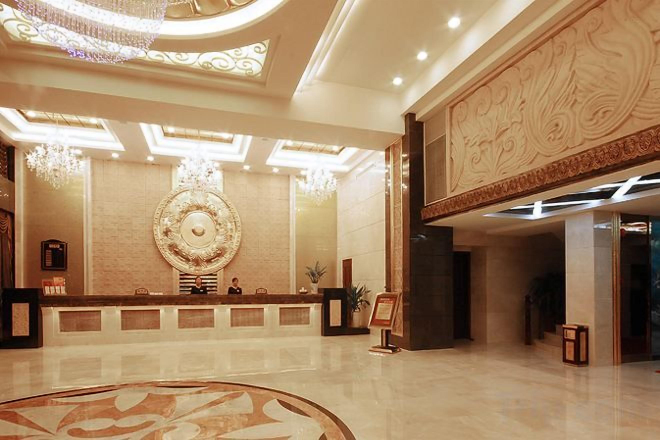Een hotel lobby met een receptiebalie, een grote decoratieve wand en een kroonluchter.