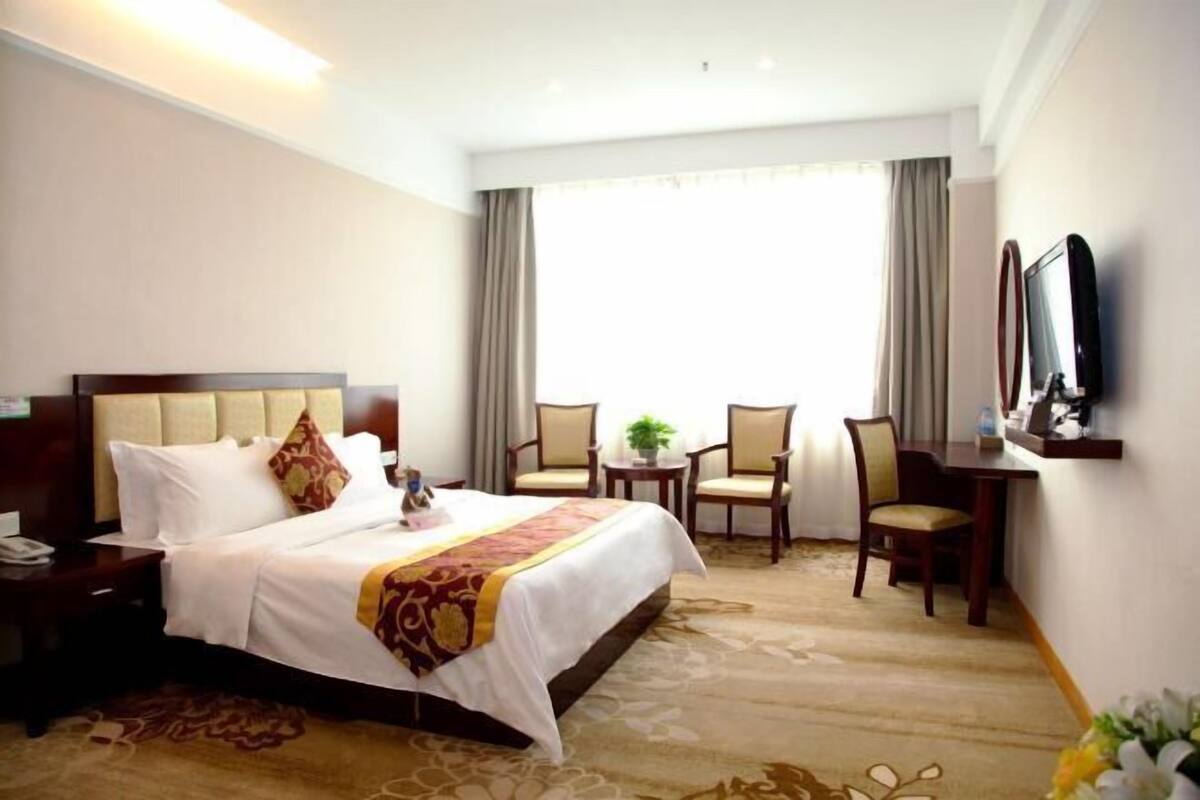 Habitación de hotel con una cama grande, un escritorio, dos sillas, un televisor y una ventana con cortinas.